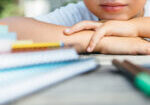 Crop shot of kid leaning head on hands while posing at table with notepads and pens.