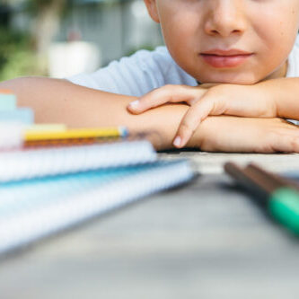 Crop shot of kid leaning head on hands while posing at table with notepads and pens.