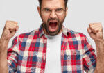 Furious angry man with irritated expression, clutches fists angrily, shouts at someone, wears checkered shirt, stands against white background. Negative human emotions and feelings. Body language