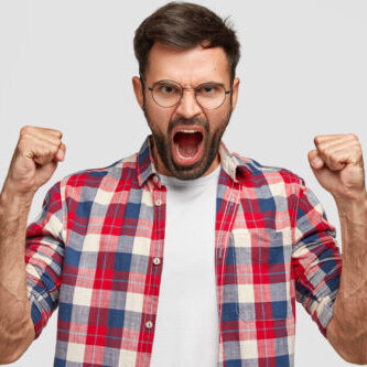 Furious angry man with irritated expression, clutches fists angrily, shouts at someone, wears checkered shirt, stands against white background. Negative human emotions and feelings. Body language