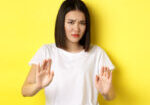 Please no. Asian woman, victim of assualt or domestic abuse, pleading with hands raised up in defense, frowning sad, begging stop, standing over yellow background.