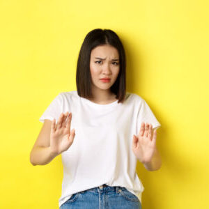 Please no. Asian woman, victim of assualt or domestic abuse, pleading with hands raised up in defense, frowning sad, begging stop, standing over yellow background.